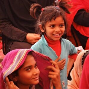 A girl with pigtails in a blue shirt smiles at something off camera, a girl with a shawl in the foreground smiles at the camera.