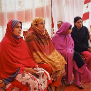 Four women in pink, red, yellow, and black shawls listen to a vocational training talk.