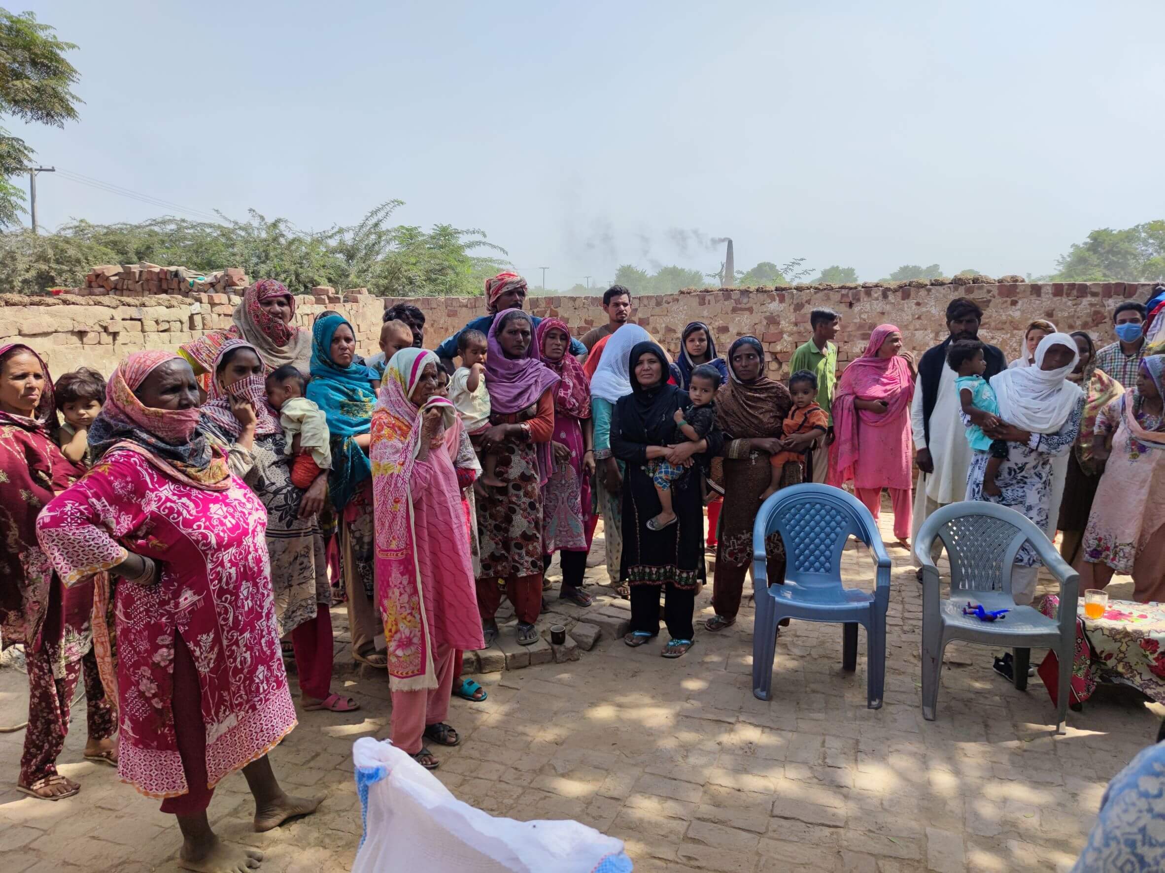 RAM Pakistan COVID Relief Distribution About 20 people stand around in the shade of tree waiting for a relief kit.