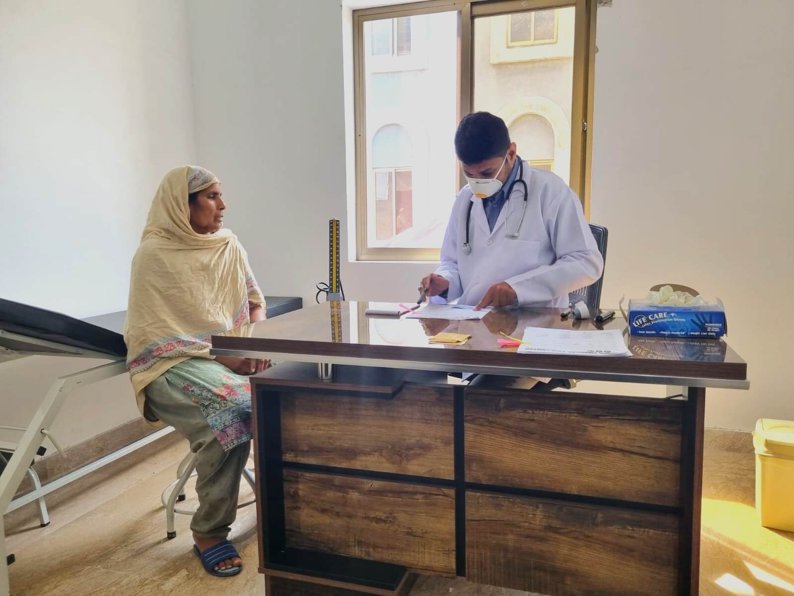 RAM Medical Clinic Patient A doctor behind a desk writes down a note for a woman in a yellow shawl sitting down next to the desk.
