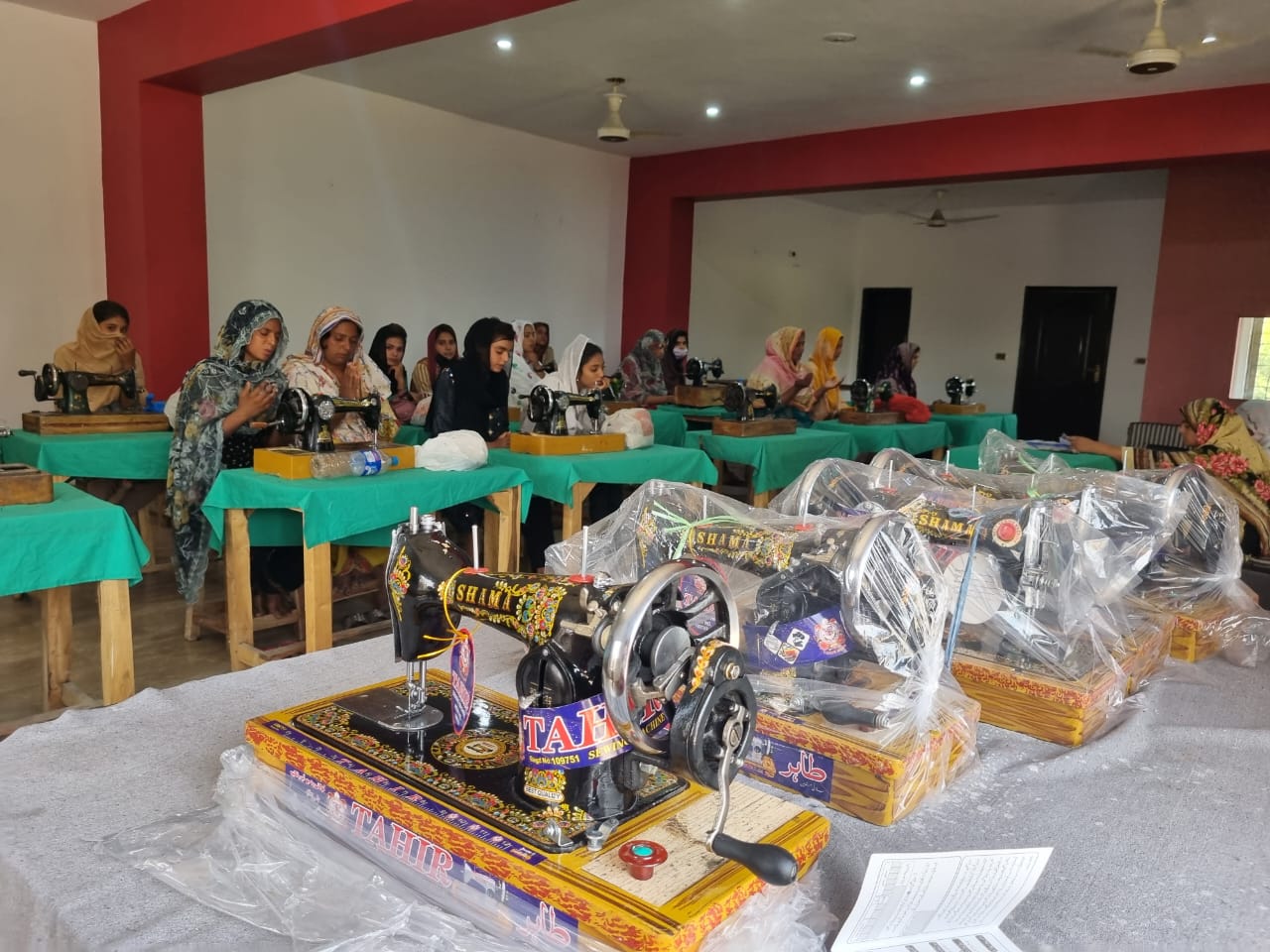 Several new sewing machines wrapped in plastic sit on a table in front of women working at small tables with sewing machines.
