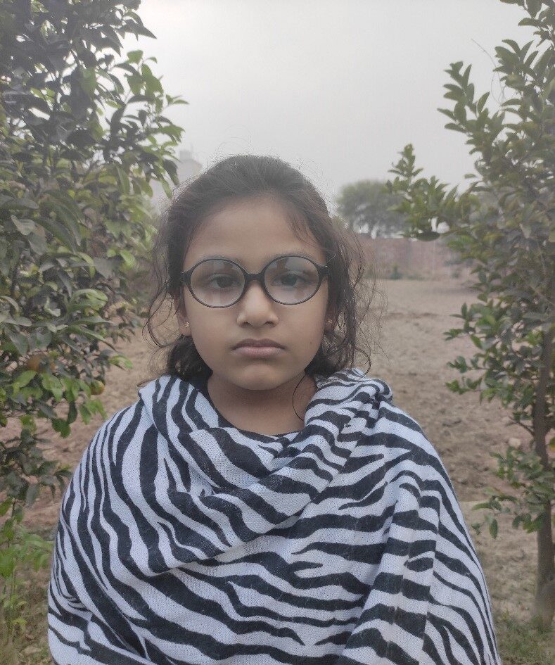 Ariela A young Pakistani girl with glasses and a zebra printed shawl stands in front of two rows of trees.