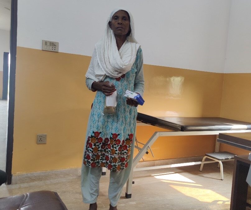 A woman in a blue shalwar kameez with a white headscarf stands in front of a table.