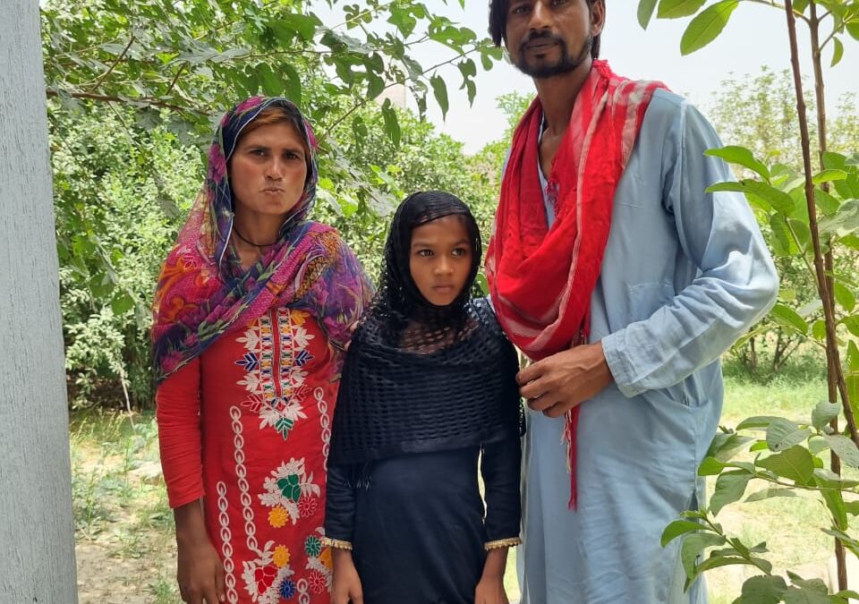 A woman in pink and white and a man in a light blue shalwar kameez stand on either side of a young girl wearing black.