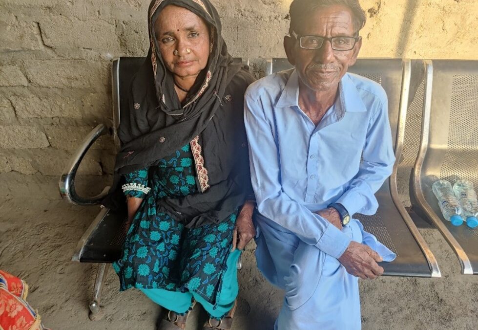 A Pakistani man in blue clothing and glasses sit's next to a woman with a black headscarf and black and turquoise clothing.