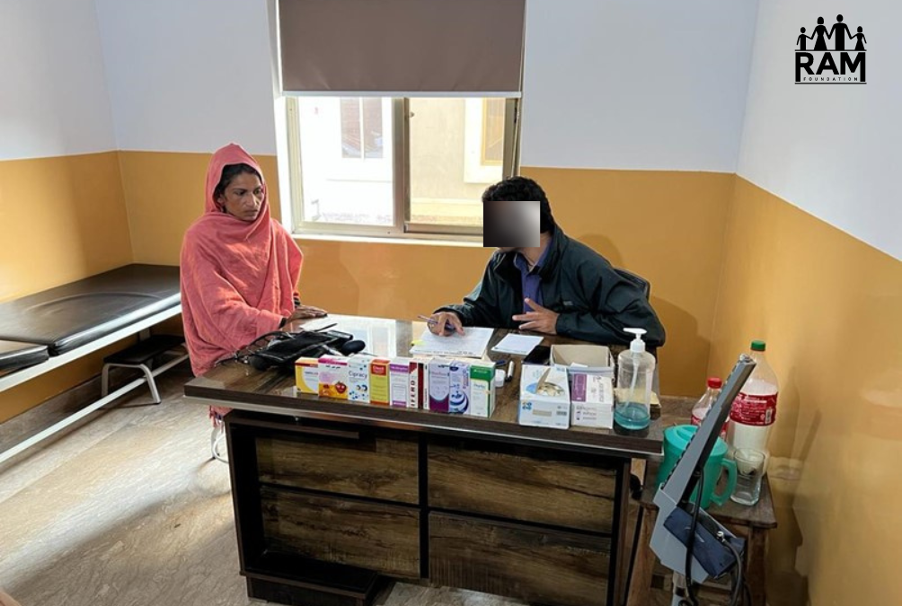 A woman in a coral head scarf sits next to desk with lots of medications on it as she talks to a doctor in black clothing.