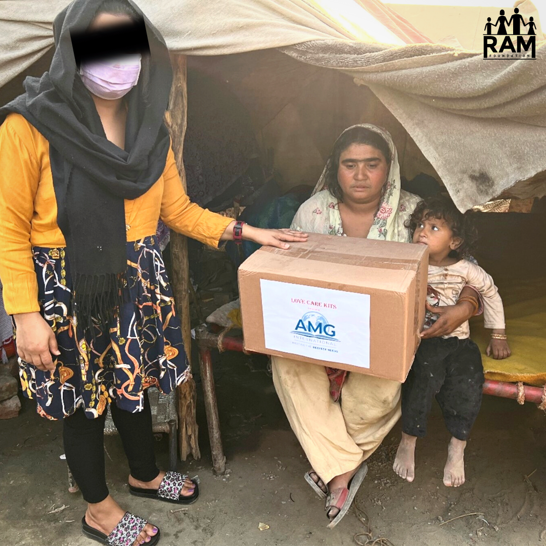 Azmat - Flood Relief - WITH LOGO A field worker in yellow and black with her face blacked out stands next to a woman in light green sitting under a makeshift tent with a box of food and hygiene items on her lap.