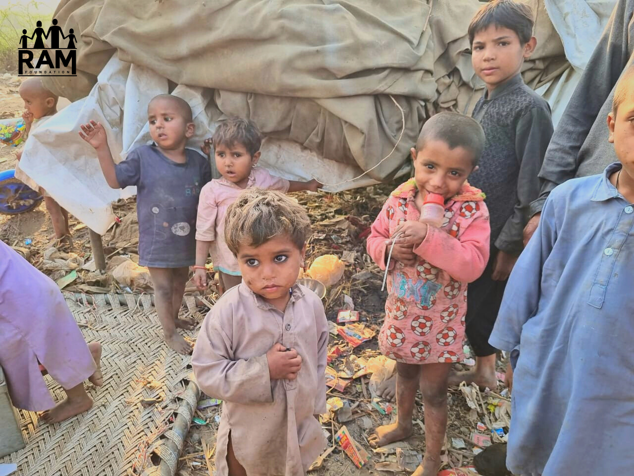 Flood Relief Round 2 - WITH LOGO EDITED Young children in dirt clothes gather round in front of a makeshift camp.