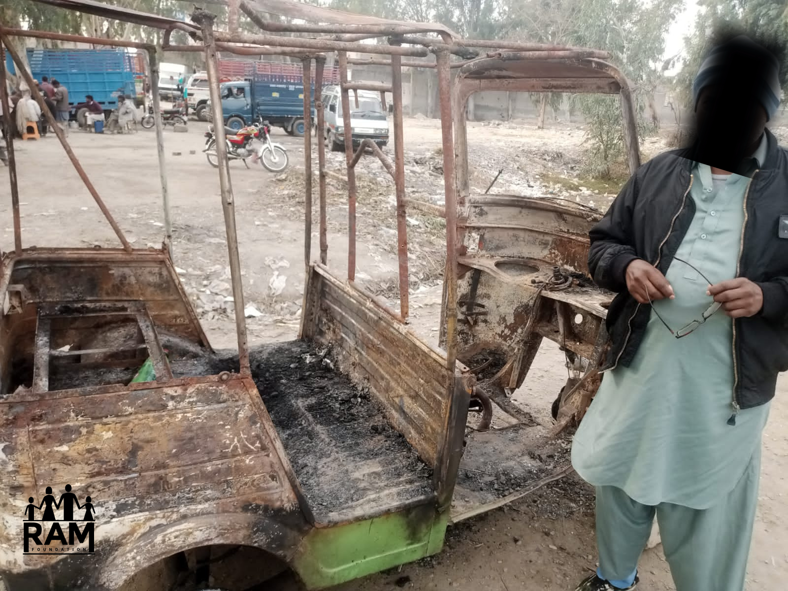 Rickshaw 1 - WITH LOGO A man with a gray shalwar kameez and his face blacked out stands next to the hollow cage of a rickshaw that was completely burnt.