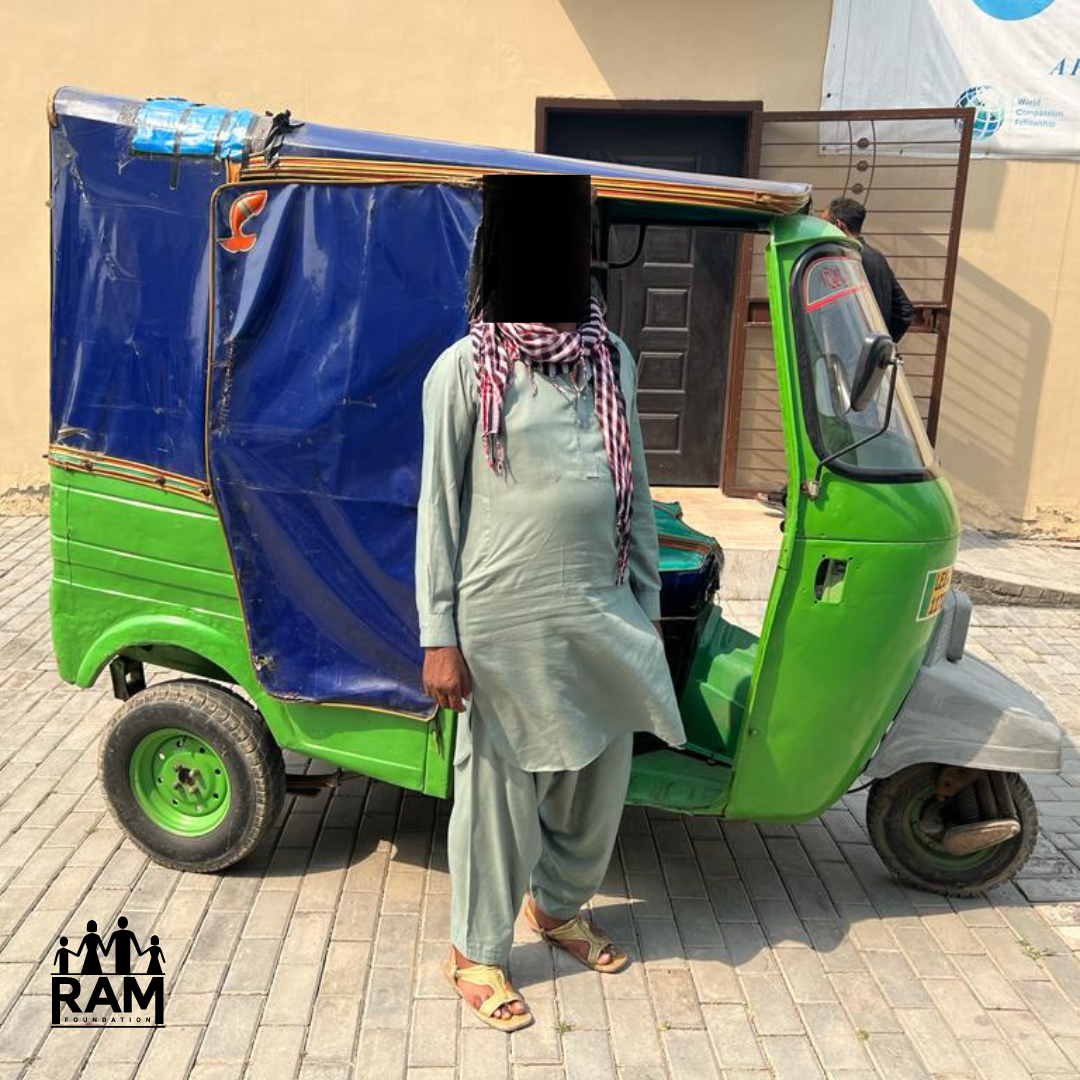 Rickshaw Burned Update - WITH LOGO A man with his face blacked out wearing a gray shalwar kameez stands in front of a green and blue rickshaw.
