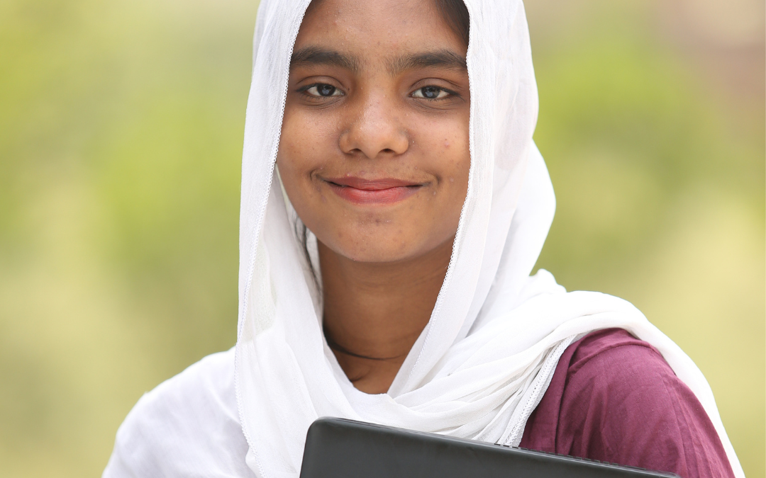 A young Pakistani woman wearing a loose white headscarf with a mauve dress smiles while holding a black laptop.